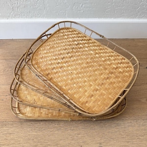 Op de afbeelding: Een stapel van drie rechthoekige geweven bamboe trays. De trays hebben afgeronde hoeken en opstaande randen, met een natuurlijke lichtbruine kleur. Het geweven patroon is zichtbaar op het oppervlak van elke tray, waardoor een textuur ontstaat.