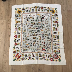 May include: A white rectangular textile featuring a detailed map of Germany with colorful illustrations of landmarks, cities, and regional coats of arms. The border is decorated with a repeating pattern of small figures in traditional clothing.