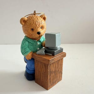 Può includere: Un ornamento a forma di orsacchiotto marrone che indossa una camicia verde abbottonata e pantaloni blu, seduto a una scrivania in legno marrone con un monitor grigio e un mouse grigio. Un filo dorato è fissato sulla testa dell'orsacchiotto.