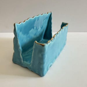 May include: A light blue ceramic napkin holder with a glossy finish and uneven, textured edges. The top rim of the rectangular structure is accented with a thin, metallic gold trim that follows the irregular contour of the ceramic walls.