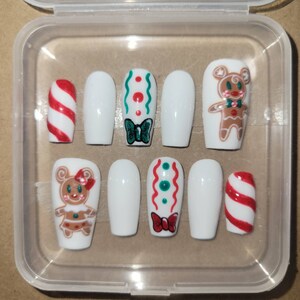 May include: A set of ten white false nails with various Christmas-themed designs. The designs include a gingerbread person, candy canes, and green and red swirls.