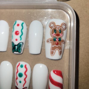 May include: Set of white false nails with Christmas-themed designs. The designs include a gingerbread man, candy canes, and a bow with red and green dots.