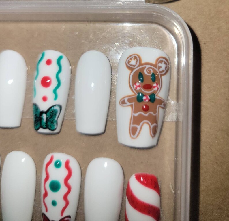 May include: Set of white false nails with Christmas-themed designs. The designs include a gingerbread man, candy canes, and a bow with red and green dots.
