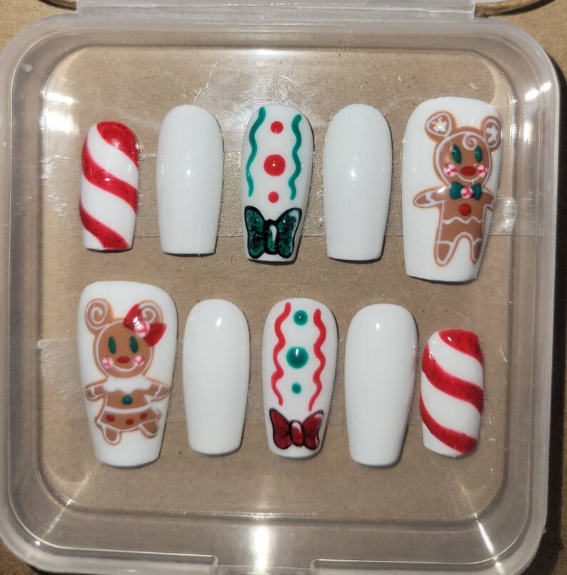 May include: Set of ten white false nails with festive Christmas designs. The designs include gingerbread people, candy canes, and bows with red and green accents.