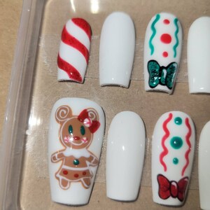 May include: Set of ten white false nails with various Christmas-themed designs. The designs include a candy cane, a green bow, a gingerbread person, and red and green swirls.