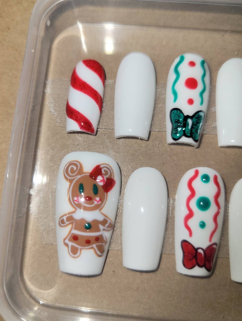 May include: Set of ten white false nails with various Christmas-themed designs. The designs include a candy cane, a green bow, a gingerbread person, and red and green swirls.