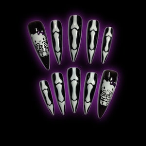 May include: A set of ten long, pointed, black press-on nails. Each nail features a white bone design or a Hello Kitty skeleton design with a purple bow. The nails are set against a dark purple and black background.