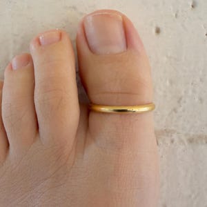 Puede incluir: Un anillo de dedo del pie dorado se usa en el dedo gordo del pie de una persona. El anillo tiene un diseño de banda simple. El tono de piel es claro y el fondo es una superficie blanca texturizada.