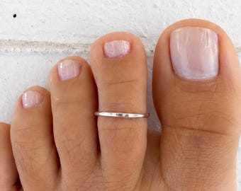 Könnte beinhalten: Ein dünner Zehenring aus poliertem, silberfarbenem Metall mit einem einfachen, glatten und kreisförmigen Banddesign. Der Ring wird am zweiten Zeh getragen und zeigt eine minimalistische Ästhetik, die mit einer metallischen Oberfläche flach aufliegt.