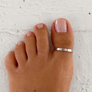 Könnte beinhalten: Ein schlichter, polierter Zehenring aus silberfarbenem Metall mit einem glatten, flachen Banddesign. Der Ring wird am großen Zeh getragen und weist eine saubere, reflektierende Oberfläche auf, die den Zeh mit gleichbleibender Breite umschließt.