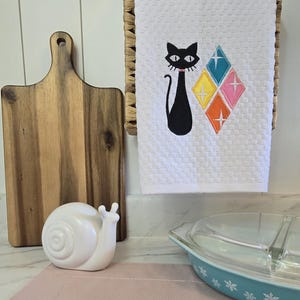May include: A white waffle-weave kitchen towel featuring an embroidered black cat with a pink collar next to four diamond-shaped icons in teal, yellow, orange, and pink, each accented with a white starburst design in the center.