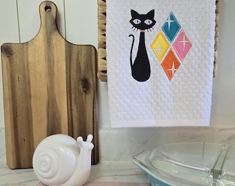 May include: A white waffle-weave kitchen towel featuring an embroidered black cat with a pink collar next to four diamond-shaped icons in teal, yellow, orange, and pink, each accented with a white starburst design in the center.