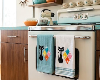 May include: Two rectangular kitchen towels hanging on a stove handle. One towel is teal and the other is white with teal stripes. Both feature a black cat silhouette next to a cluster of four colorful diamond shapes with starburst accents.