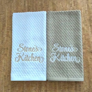 May include: Two kitchen towels with a textured pattern. One is white with gold embroidery, the other is tan with white embroidery. Both towels have the text "Stone's Kitchen" embroidered on them.