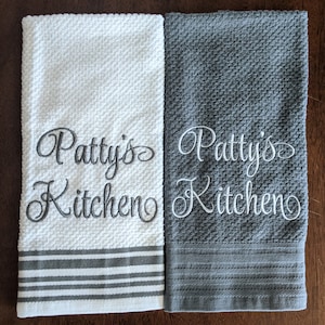 May include: Two kitchen towels with gray and white stripes. One towel is white with gray embroidery that says "Patty's Kitchen". The other towel is gray with white embroidery that says "Patty's Kitchen".