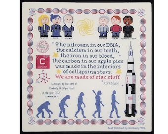 May include: A cross-stitch embroidery featuring a quote by Carl Sagan about the origins of the elements in our bodies. The quote reads "The nitrogen in our DNA, the calcium in our teeth, the iron in our blood, the carbon in our apple pies was made in the interiors of collapsing stars. We are made of star stuff." The embroidery also includes a depiction of a rocket, a DNA strand, and a series of stick figures representing the evolution of humans.