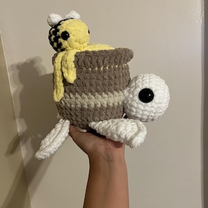 May include: A handmade crochet turtle with a honey pot shell and a bee on top. The turtle is white with a large black eye. The honey pot is brown and yellow, with a bee peeking out. The bee is yellow and black.