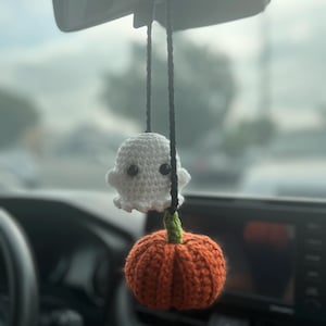 May include: A handmade crochet car charm featuring a white ghost and an orange pumpkin. The ghost has black button eyes and a friendly expression. The pumpkin has a green stem. The charm hangs from a black string, perfect for Halloween.
