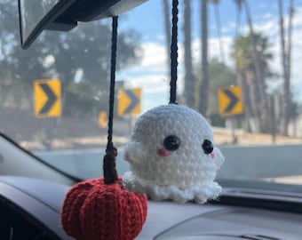 May include: A crocheted white ghost with black eyes and pink cheeks hanging next to a crocheted red pumpkin. Both items are attached to black braided cords suspended from a vehicle rearview mirror, featuring textured yarn construction.