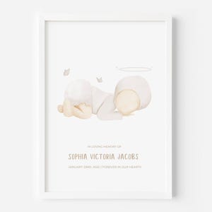 Puede incluir: Una impresión enmarcada en blanco con una ilustración en acuarela de un bebé con gorro y pañal blancos, acostado con un halo sobre la cabeza y dos pequeñas mariposas. El texto dice: IN LOVING MEMORY OF SOPHIA VICTORIA JACOBS JANUARY 23RD, 2022 | FOREVER IN OUR HEARTS.