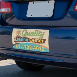May include: A blue car's rear bumper with a rectangular magnet. The magnet has the words "Quality Learing Center" and a phone number. The car is parked in a parking lot.