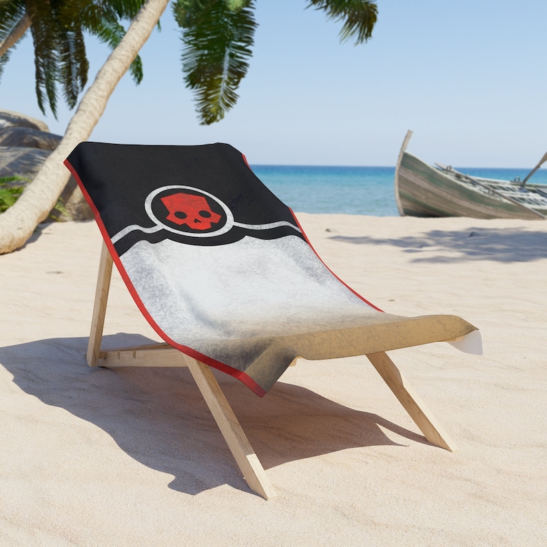 Puede incluir: Una toalla de playa negra y blanca con un dise&ntilde;o de calavera y huesos cruzados rojos en una silla de playa de madera. La silla est&aacute; en una playa de arena con palmeras en el fondo.