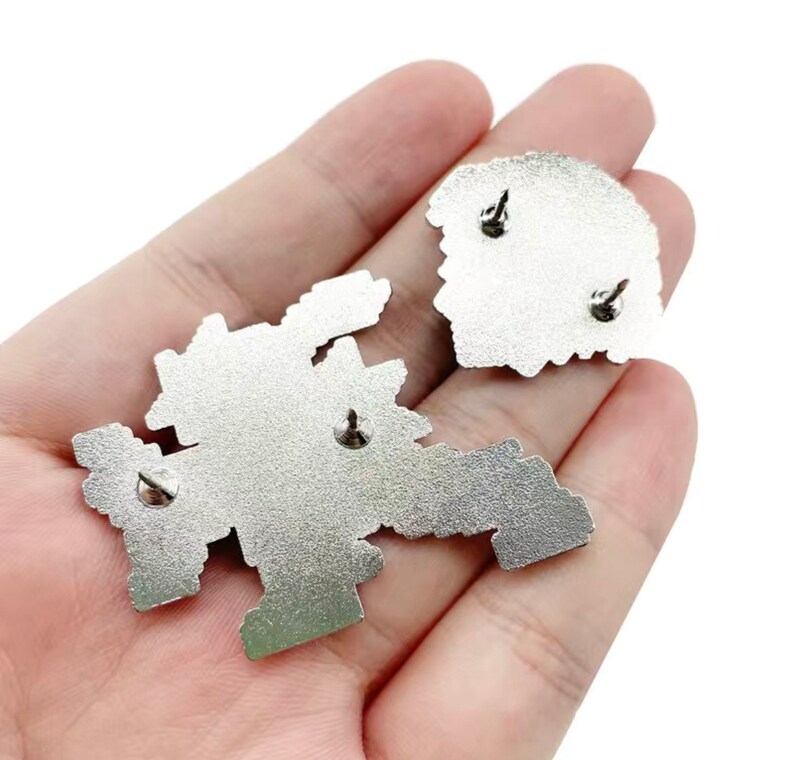 May include: Two silver metal enamel pins with a pixelated design. The pins have a safety clasp on the back.