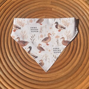 May include: White triangular pet bandana with a duck and floral pattern. The bandana features illustrations of ducks in various poses, along with the text "ZERO DUCKS GIVEN" in a serif font. The background includes floral and plant elements.