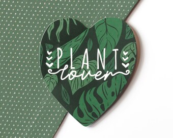 May include: A heart-shaped sticker with a dark green background featuring a pattern of tropical leaves. The words "PLANT lover" are written in white script across the center. The sticker is placed on a green dotted background.