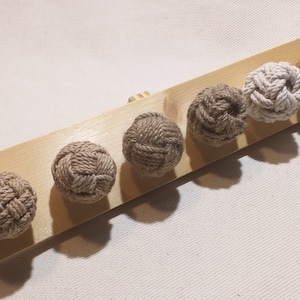 Può includere: Sei nodi decorativi in corda di diverse tonalità di marrone e bianco sono disposti su una superficie di legno. I nodi sono disposti in fila, con il nodo più grande a destra e il più piccolo a sinistra.