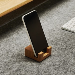 Op de afbeelding: Een moderne bureau-opstelling met een houten monitorstandaard, een smartphone in een houten houder, een draadloos toetsenbord en een muis. Een roze notitieboek ligt op de standaard. Het bureaublad is bedekt met een grijze vilten mat.