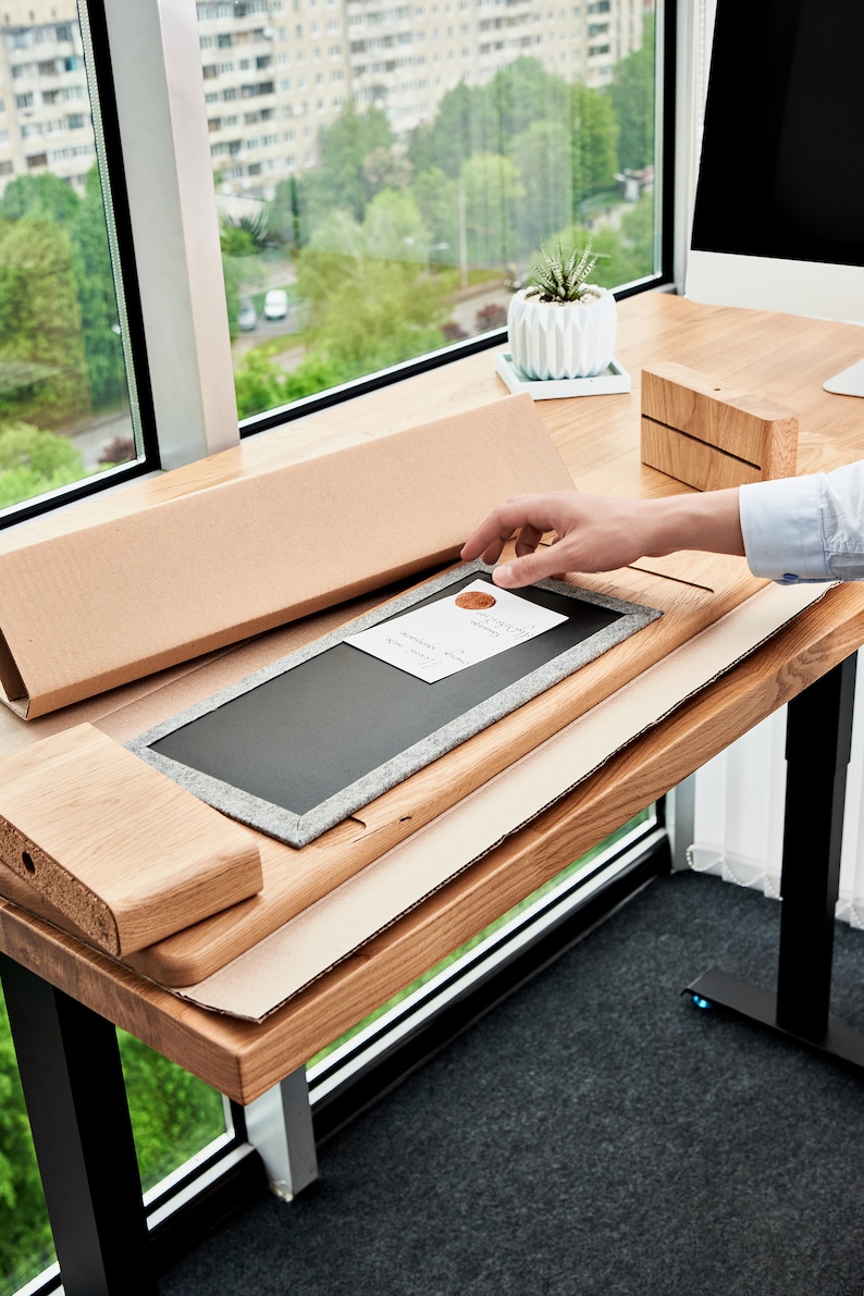 May include: A person assembling a wooden desk with a black chalkboard insert. The desk is made of light wood and has a black metal frame. The person is holding the chalkboard insert in their hand.