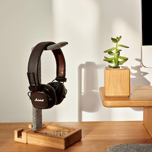 Puede incluir: Auriculares Marshall de cuero marrón sobre un soporte de madera con una pequeña bandeja con clips. Una pequeña planta suculenta en una maceta cúbica de madera se encuentra en un estante de madera. La escena está ambientada en un escritorio de madera.