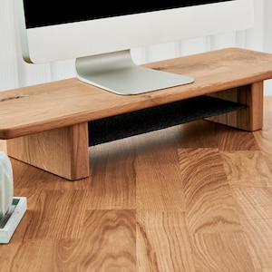May include: A wooden monitor stand with a black shelf underneath. The stand is made of light brown wood and has a natural finish. The stand is sitting on a wooden desk.