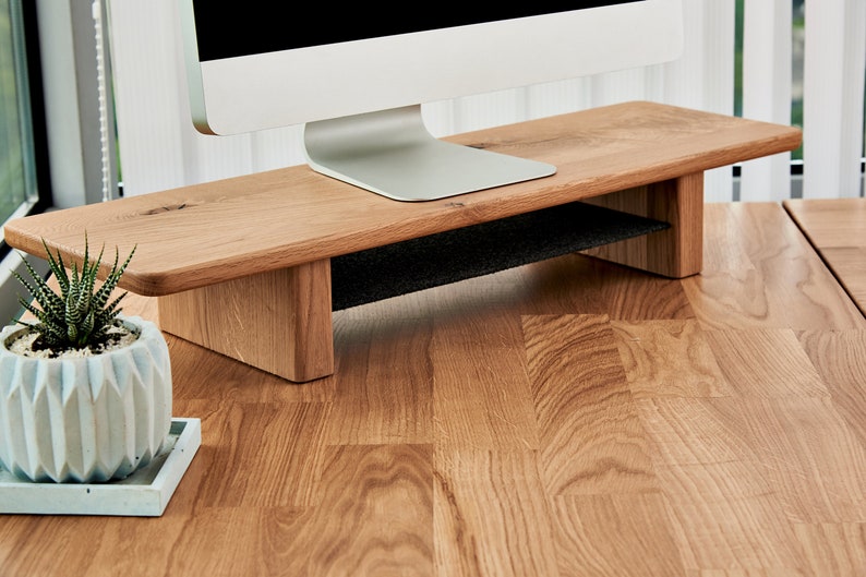 May include: A wooden monitor stand with a black shelf underneath. The stand is made of light brown wood and has a natural finish. The stand is sitting on a wooden desk.