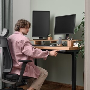 May include: A person sitting in a black office chair with a pink shirt and beige pants, adjusting the height of a black standing desk with a wooden top. Two black computer monitors are on the desk, along with a smartphone, pencils, and a small potted plant.