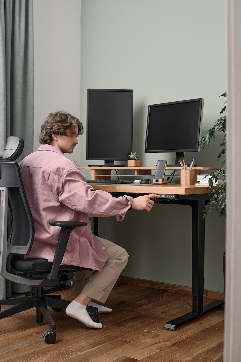 May include: A person sitting in a black office chair with a pink shirt and beige pants, adjusting the height of a black standing desk with a wooden top. Two black computer monitors are on the desk, along with a smartphone, pencils, and a small potted plant.