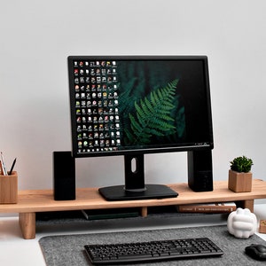 May include: A black computer monitor with a green fern wallpaper sits on a wooden monitor stand. The stand has a lower shelf for a keyboard and other items. There is a small potted plant, a pencil holder, and a white sculpture on the desk.