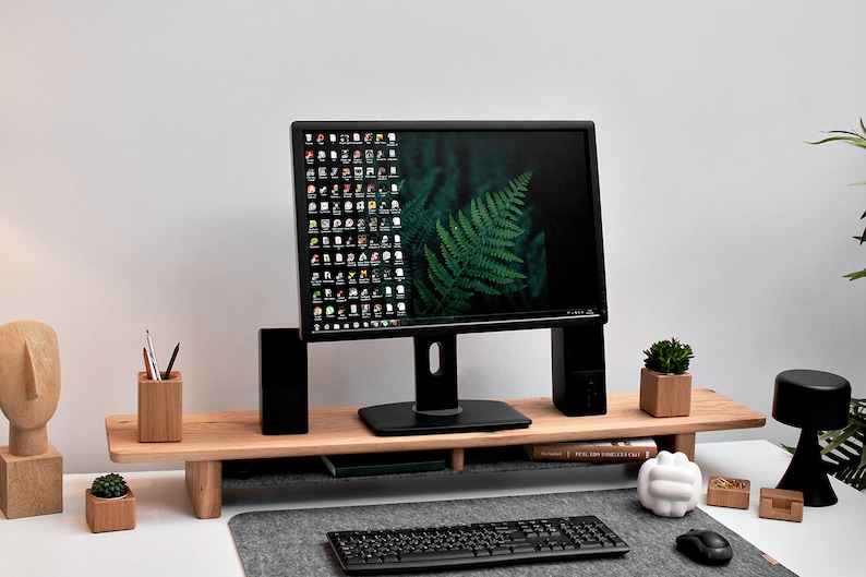 May include: A black computer monitor with a green fern wallpaper sits on a wooden monitor stand. The stand has a lower shelf for a keyboard and other items. There is a small potted plant, a pencil holder, and a white sculpture on the desk.