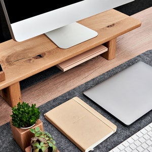 May include: A wooden monitor stand with a phone holder, a book, and two small potted plants. The stand is on a desk with a laptop, a keyboard, and a gray felt desk mat.