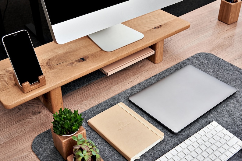 May include: A wooden monitor stand with a phone holder, a book, and two small potted plants. The stand is on a desk with a laptop, a keyboard, and a gray felt desk mat.