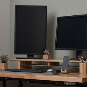 May include: Two black computer monitors are mounted on a wooden stand with a keyboard, mouse, and phone holder. The stand is on a light wood desk with a potted plant on the left and a plant on the right.