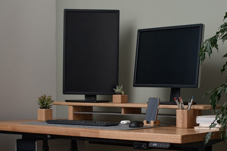 May include: Two black computer monitors are mounted on a wooden stand with a keyboard, mouse, and phone holder. The stand is on a light wood desk with a potted plant on the left and a plant on the right.