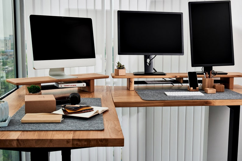 May include: Two computer monitors on wooden risers on a light brown wooden desk. The desk has a gray felt desk mat. The monitor on the left is a white iMac with a black screen. The monitor on the right is a black monitor with a black screen. There are various office supplies on the desk, including a small potted plant, a book, a pair of headphones, a phone, and a pencil holder.