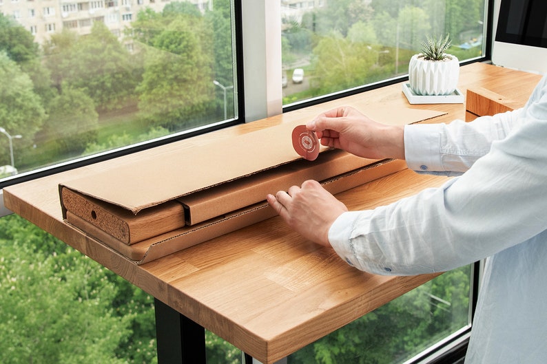 May include: A person assembling a wooden table from a flat-pack box. The table is made of light wood and has a natural finish. The person is using their hands to put the table together. The table is on a wooden surface.
