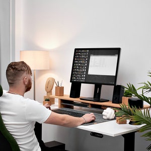 May include: A person sits at a white desk with a black computer monitor, a wooden keyboard tray, and a green office chair. The desk has a black and gray mouse pad and a white knot-shaped object. A potted plant is on the right side of the desk.