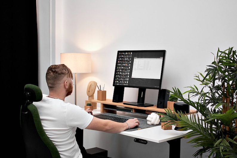 May include: A person sits at a white desk with a black computer monitor, a wooden keyboard tray, and a green office chair. The desk has a black and gray mouse pad and a white knot-shaped object. A potted plant is on the right side of the desk.