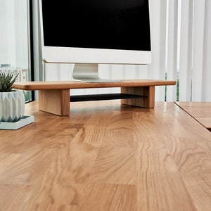 May include: A wooden computer monitor stand with a black mesh shelf. The stand is on a light brown wooden desk.