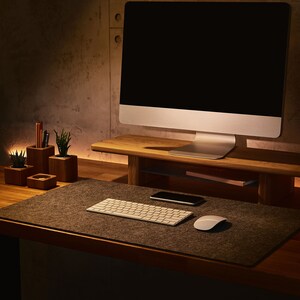May include: A modern workspace with a computer monitor on a wooden stand, a keyboard, a mouse, and a phone on a gray felt desk mat. There are also three small wooden containers with plants and pencils on the desk.