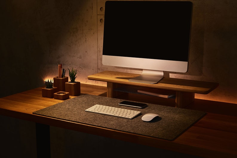 May include: A modern workspace with a computer monitor on a wooden stand, a keyboard, a mouse, and a phone on a gray felt desk mat. There are also three small wooden containers with plants and pencils on the desk.
